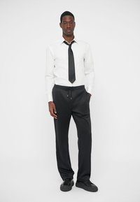 White button-up shirt with long sleeves, black tie, and black drawstring sweatpants. Black sneakers complete the outfit. Simple and modern design.