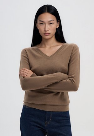 Donna con capelli lunghi e neri che indossa un maglione marrone a scollo a V e jeans scuri, in piedi con le braccia incrociate davanti a uno sfondo chiaro e semplice.