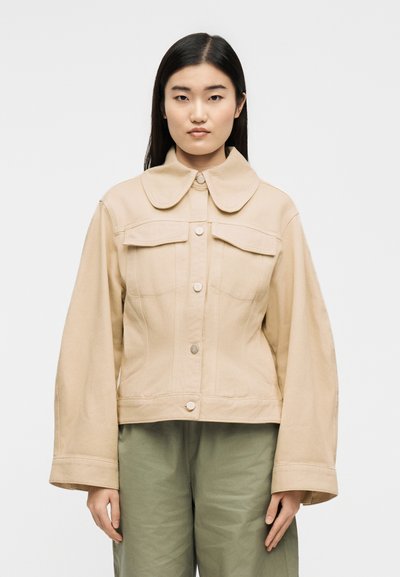 Γυναίκα με ίσια μαύρα μαλλιά που φοράει μια μπεζ φαρδιά ζακέτα με μεγάλο γιακά και χακί παντελόνι, στέκεται μπροστά σε απλό φόντο.