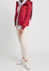 Chaqueta roja con patrón y capucha, combinada con una sudadera gris a rayas y pantalones deportivos beige. Zapatillas blancas completan el atuendo.