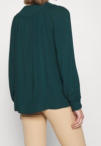 Blouse vert foncé en tissu doux, avec un dos plissé, des poignets boutonnés et un col classique. Portée par-dessus un pantalon beige clair ajusté.