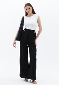 Witte mouwloze top, zwarte hoge taille broek met wijd uitlopende pijpen en gouden hardware, en metallic gouden sandalen met banden. Zwarte schoudertas.