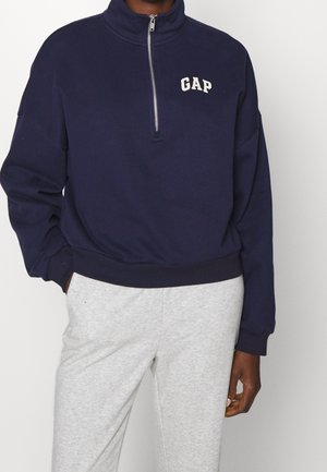 Personne portant un sweat GAP bleu marine à demi-fermeture éclair et un pantalon de survêtement gris clair, debout avec une main dans la poche sur un fond uni.
