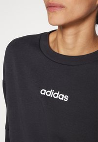 Fekete pulóver kerek nyakkivágással, sima textúrával, a mellkasán feltűnő fehér "adidas" logóval.