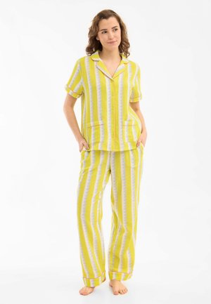 Woman standing barefoot wearing yellow and white striped button-up short sleeve top with matching loose pants, hands in pockets, looking sideways.