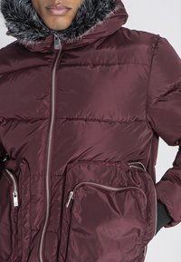 Maroonowa kurtka puchowa z odpinanym sztucznym futrem, dwoma przednimi kieszeniami na zamek błyskawiczny oraz gładką, błyszczącą teksturą. Posiada centralny zamek.