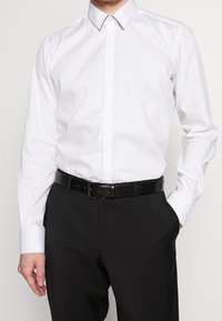 Biała koszula z długim rękawem i klasycznym kołnierzykiem, o subtelnym połysku. Do tego czarny, teksturowany skórzany pasek, noszony z ciemnymi spodniami.