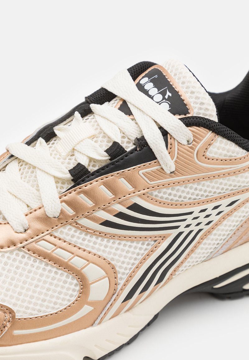Sneaker met een bovenwerk van mesh in crème, zwart en metallic goudaccenten, met een textuuroverlaag en crèmekleurige veters op een lichtgewicht zool.
