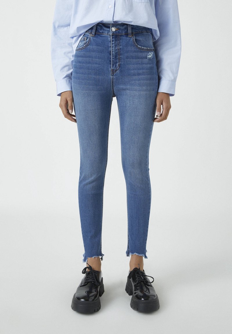 Vaqueros de mezclilla azul ajustados con una cintura alta, que presentan un dobladillo deshilachado y un sutil desgaste. Combinados con zapatos de plataforma negros.