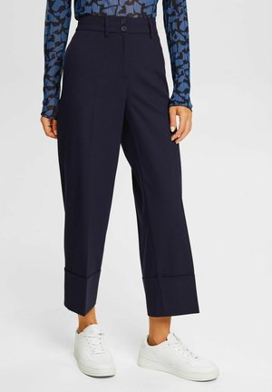 Vrouw die donkere marineblauwe cropped wijde broek met omgeslagen zomen draagt, een blauwe lange mouwen top met patroon en witte sneakers.