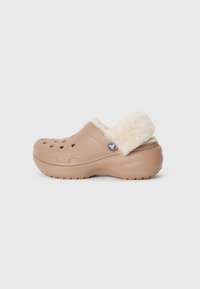 Zoccolo beige con fodera cremosa e pelosa, fori ventilati sulla parte superiore e suola texturizzata per una presa sicura. Presenta un'accattivante accentuazione metallica decorativa.