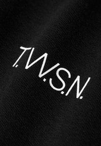 Zwarte stof met een gestructureerd oppervlak waarop de witte tekst "T.W.S.N." staat in een modern, schreefloos lettertype. Duidelijke en vetgedrukte letters.
