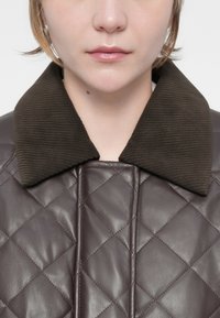 Gequilte chocoladebruin leren jas met een gestructureerde donkergroene corduroy kraag, voorzien van ruitpatronen en contrasterende stiksels.