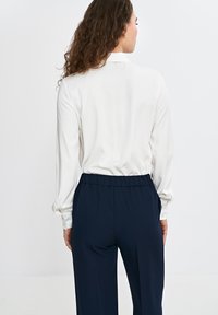 Lekka, biała bluzka z długim rękawem, z mankietami na guziki i wysokim kołnierzem, w połączeniu z granatowymi spodniami elastycznymi w pasie o gładkiej teksturze.