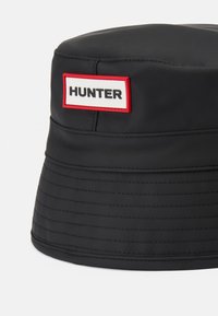 Čierny gumový vedierkový klobúk s ribbenovou textúrou. Obsahuje bielu nálepku s červeným okrajom a výrazným textom "HUNTER" na prednej strane.