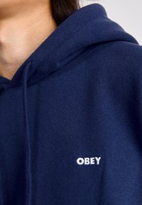 Sweatshirt azul marinho feito de tecido macio, com um bolso frontal e um logo "OBEY" bordado em branco no peito. Capuz com cordão ajustável.