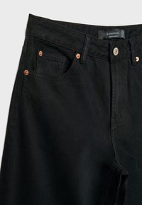 Gros plan sur la zone de la taille d’un jean noir en denim avec des rivets en cuivre, une fermeture par bouton, des poches avant, et une étiquette de marque visible à l’intérieur de la ceinture.