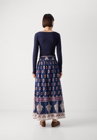 Haut à manches longues bleu marine associé à une jupe maxi à motifs floraux en bleu, blanc et rouge, avec des bordures décoratives et une ceinture élastique.