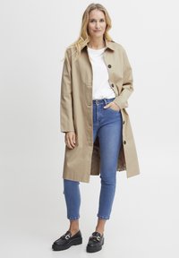 Beiger Trenchcoat mit lässiger Passform, großen Frontknöpfen und seitlichen Taschen, kombiniert mit blauen Jeans und schwarzen Loafers.