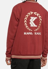 Persona che indossa una giacca color bordeaux con polsini e cintura a strisce beige, decorata con una foglia di alloro bianca e il logo "KARL KANI" sul retro.