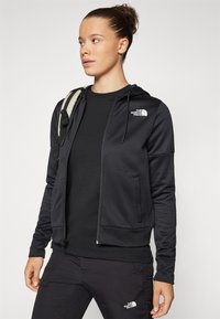 Black zip-up hoodie with a soft texture, featuring a small white logo on the chest, paired with a black t-shirt and matching pants.