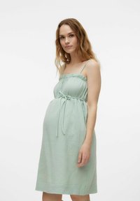 Miętowa sukienka ciążowa z teksturowanej tkaniny, z cienkimi ramiączkami, falbaną przy dekolcie i paskiem w talii do regulacji dopasowania.