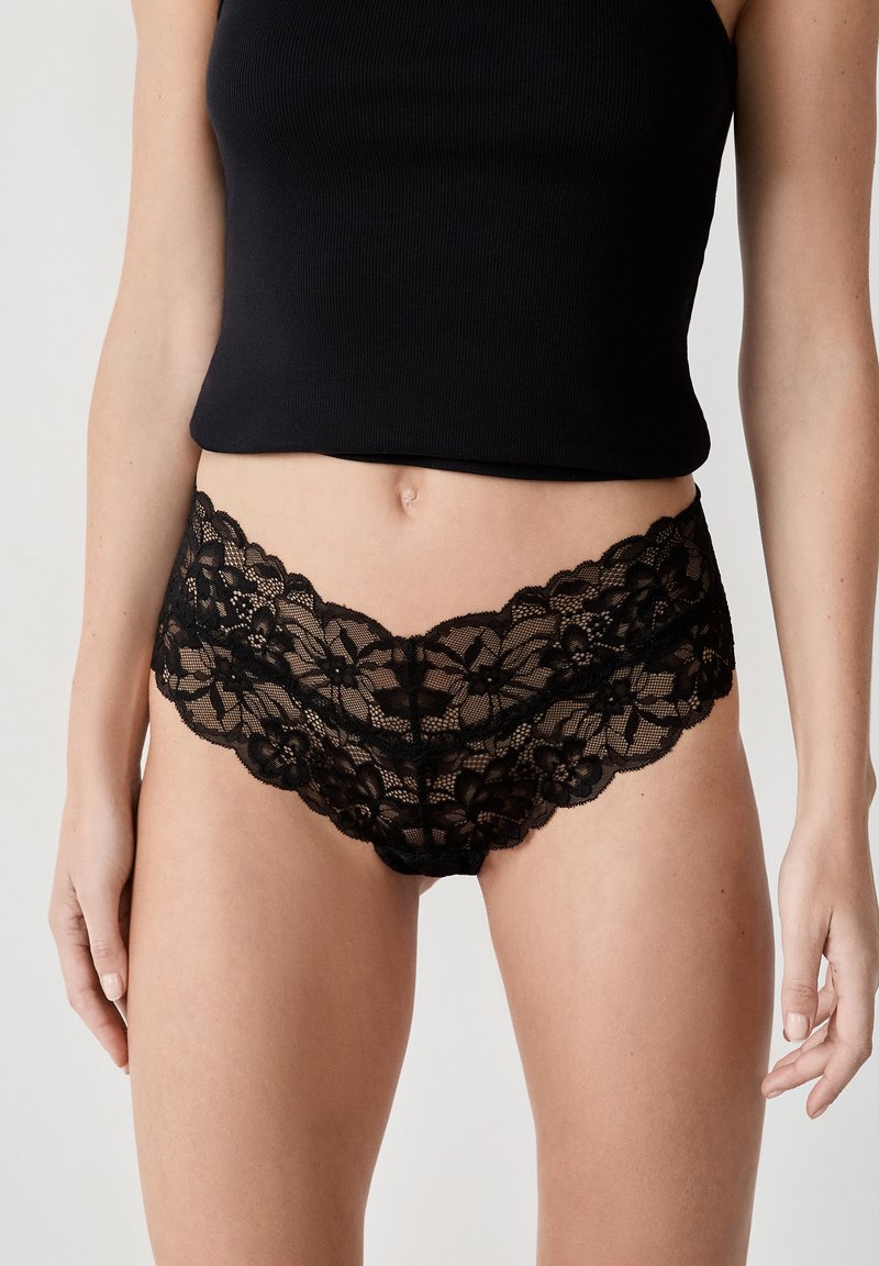 Pantie nere di pizzo con motivo floreale e bordi ondulati, caratterizzate da una texture morbida e un design a vita bassa, abbinate a un top nero.