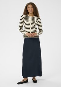 Ženska s kodrasto obleko, ki nosi bež in črno prugasto z gumbi v blagovni obliki, dolgo mornarsko krilo in črne balerinke, stoji z rokami sklenjenimi.