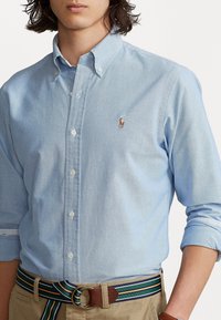 Jasnoniebieska koszula z guzikami i małym logo, w zestawieniu z beżowym paskiem w paski w odcieniach zieleni, niebieskiego i beżu. Gładka tekstura materiału.