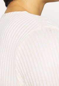 Top de color beige claro con canales en relieve que presenta finas rayas verticales, textura suave y un clásico escote redondo, con un ajuste relajado en la zona de los hombros.