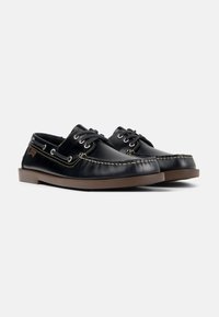 Zapatos de barco de cuero negro con un diseño cosido, ojales de metal, suela de goma marrón y un contraste de costuras en tono claro a lo largo de la parte superior.
