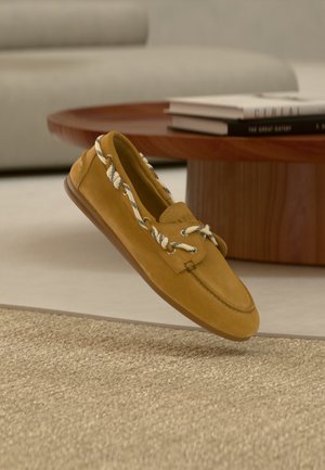 Tan suede loafer with white and black rope laces, positioned above a beige woven rug beside a wooden coffee table with books.