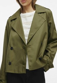 Olive krāsas dubulto pogu žakete, izgatavota no gludas auduma, ar lieliem melniem pogām un plašu apkakli, valkāta virs balta adīta blūze.