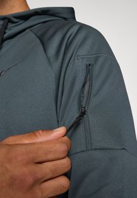 Nike-jakke med mørkegrått stoff, en glidelåslomme på ermet og en glatt, strukturert overflate med et tettsittende design.