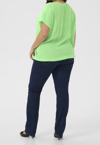 Personne portant un haut à manches courtes vert vif, un jean bleu foncé et des chaussures noires à talons, le dos légèrement tourné.