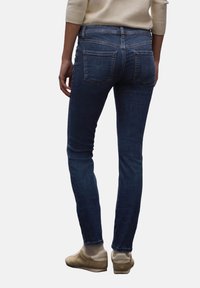 Jeans de mezclilla ajustados en un lavado oscuro, con cinco bolsillos, un ligero desvanecido y una cinturilla de altura media. Combinados con zapatillas beige.