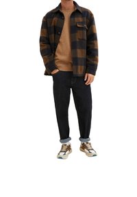 Mann in braun-schwarz kariertem Sakko, beiger Strickjacke, dunkelgerollten Jeans und braunen, weißen und blauen Sneakers, steht mit einer Hand in der Tasche.