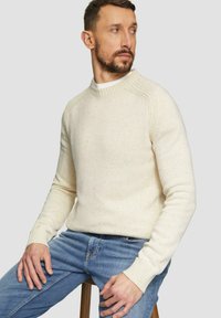 Sweter w kolorze kremowym z dzianiny, z ściągaczami przy mankietach i dekolcie, charakteryzujący się teksturowanym wzorem i przylegającym krojem. Noszony z niebieskimi dżinsami.