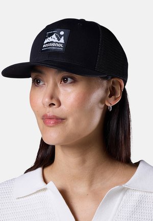 Frau trägt eine schwarze Rossignol-Mütze mit Berglogo, weißes Strickshirt und kleine Creolen, schaut leicht nach rechts vor weißem Hintergrund.