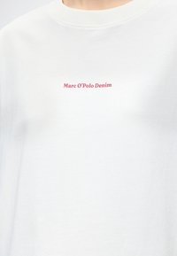 Λευκό μπλουζάκι με στρογγυλή λαιμόκοψη και μικρό κόκκινο κείμενο "Marc O’Polo Denim" στο κέντρο στο στήθος.