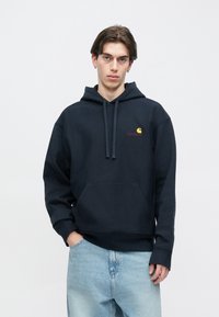 Μπλε ναυτικό φούτερ από βαμβακερό μείγμα με τσέπη καγκουρό, ρυθμιζόμενο κορδόνι και λογότυπο Carhartt σε κίτρινο και κόκκινο στο στήθος.