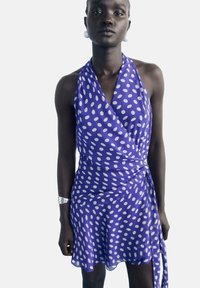 Femeie îmbrăcată într-o rochie wrap cu guler halter de culoare violet cu buline albe, purtând o brățară din argint și cercei mari din perlă albă, pe un fundal simplu.