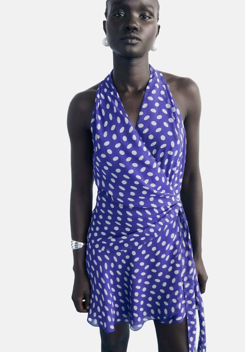 Femeie îmbrăcată într-o rochie wrap cu guler halter de culoare violet cu buline albe, purtând o brățară din argint și cercei mari din perlă albă, pe un fundal simplu.