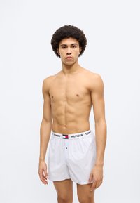 Άσπρα ριγέ μποξεράκια με μαύρη και κόκκινη λωρίδα μέσης με λογότυπο, που διαθέτουν κλείσιμο με κουμπί μπροστά. Το μοντέλο παρουσιάζει το προϊόν.