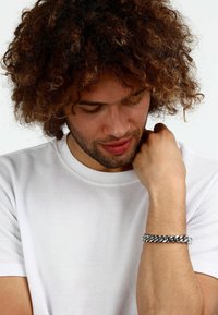 Bracciale in metallo argentato con catena sul polso, caratterizzato da anelli intrecciati, in contrasto con una camicia bianca a trama con scollo rotondo.