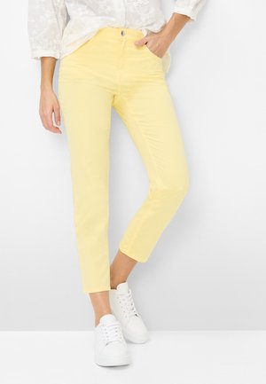 Kvinde iført lysegule, tætsiddende bukser, hvid bluse med blomstermønster og hvide sneakers, stående med den ene hånd i lommen mod hvid baggrund.