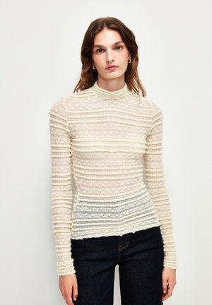 Langarmshirt in Creme mit strukturierten Streifen, hochgeschlossen und figurbetont. Der Saum verläuft in ein helles Blau und erzeugt einen Farbverlaufseffekt.