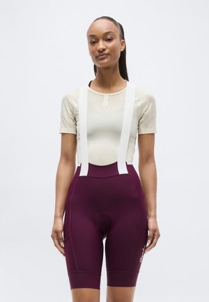 Jeune femme portant un haut beige clair à manches courtes et un short de cyclisme violet taille haute avec des bretelles blanches, debout devant un fond uni.