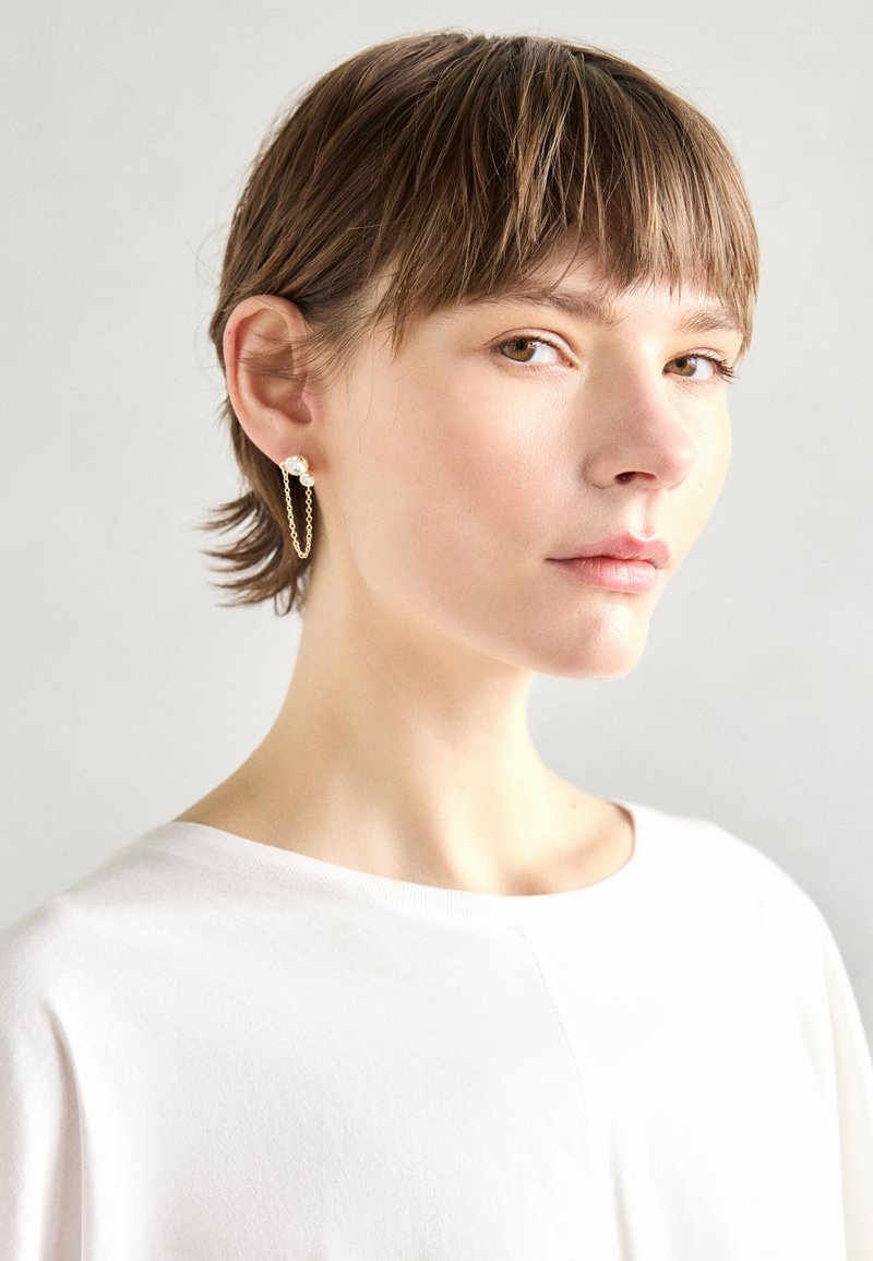 Pendientes de cadena de oro con una perla decorativa, con un diseño minimalista. El modelo lleva una camisa blanca sencilla, mostrando la joyería.