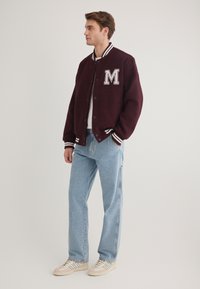 Burgunderfarbene Collegejacke mit grauem "M"-Emblem, weißen gestreiften Bündchen und Kragen, kombiniert mit hellblauen schmal geschnittenen Jeans und beigen Sneakers.
